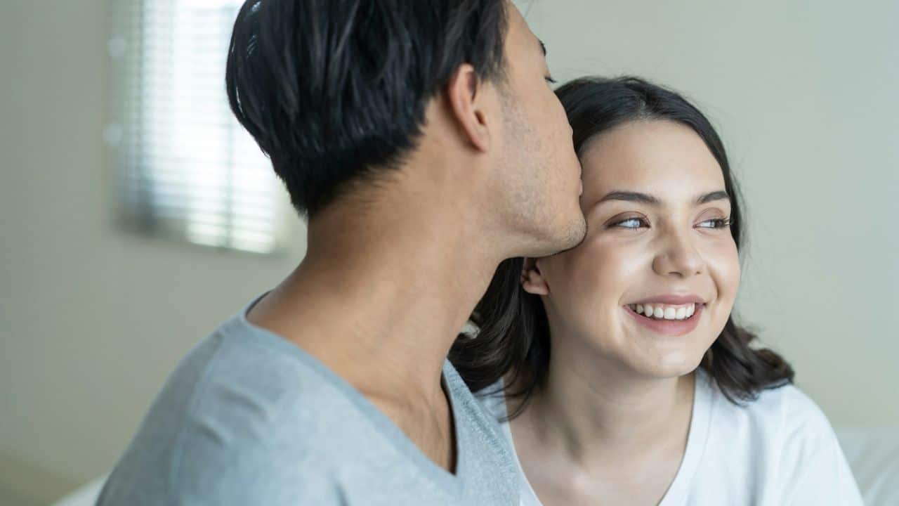 A man is kissing a smiling woman on her cheek in a bright, indoor setting.