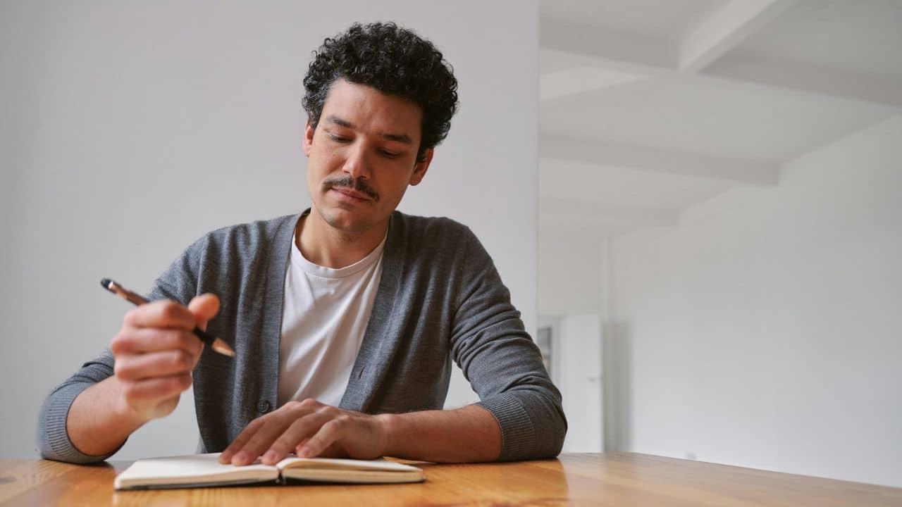 A man with dark curly hair and a mustache writing in a notebook on a wooden table.