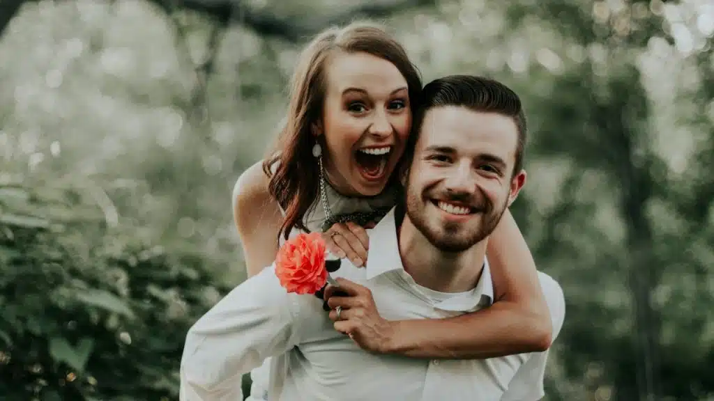 A happy woman holding a flower in her hand riding on the back of a man.