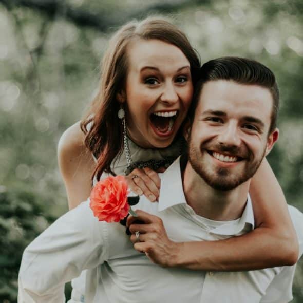 A happy woman holding a flower in her hand riding on the back of a man.