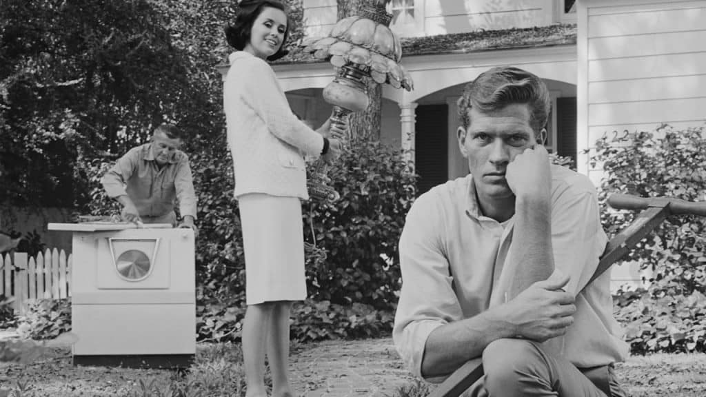 A bored man kneeling down next to a wheelbarrow while a woman standing behind is looking at him.