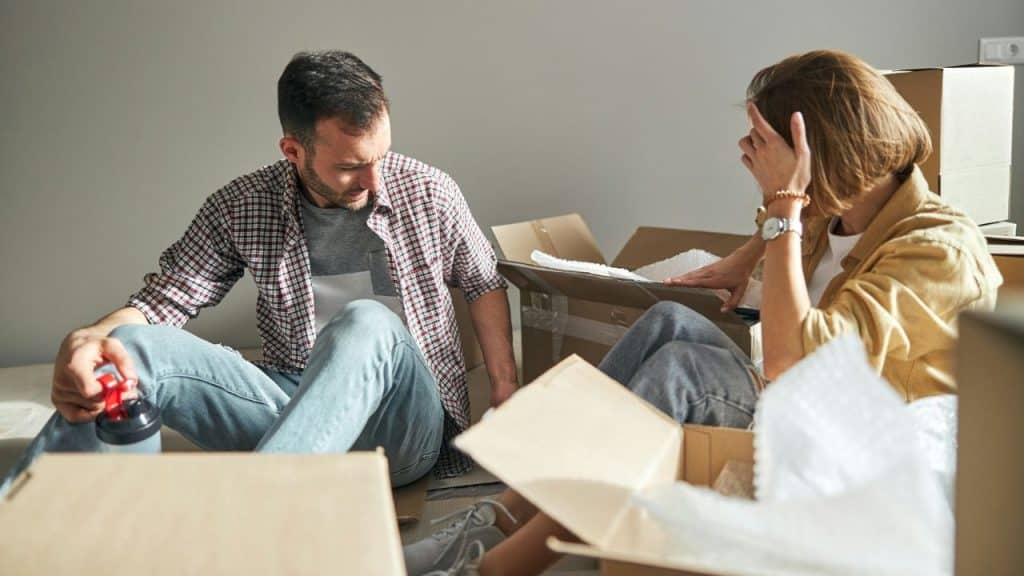 A man and a woman sit on the floor surrounded by open moving boxes.