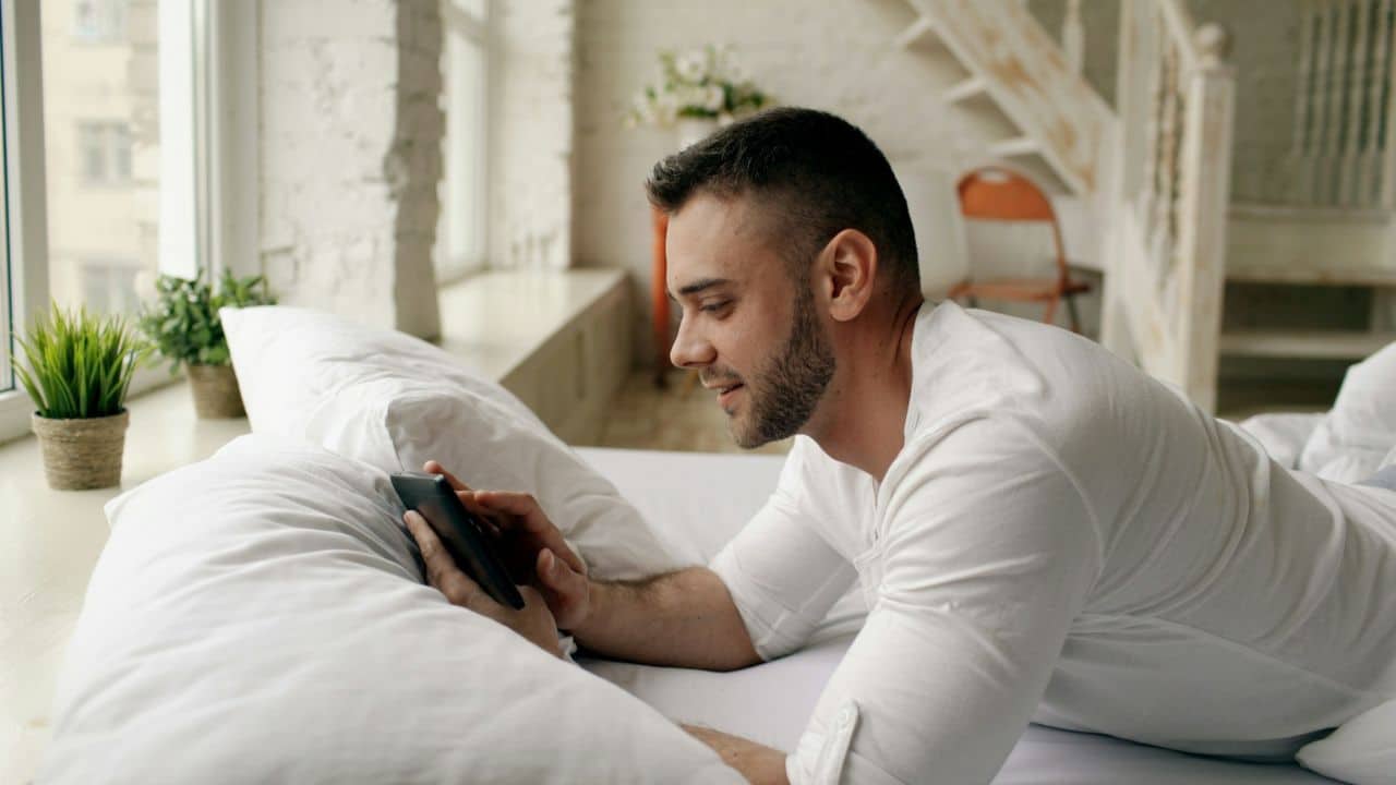A man in a white shirt lies in bed looking at his phone near a bright window.