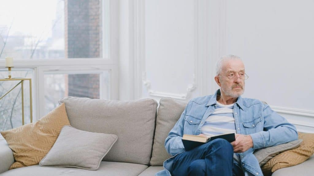 A senior man sitting on a sofa holding a book and looking thoughtfully out the window.