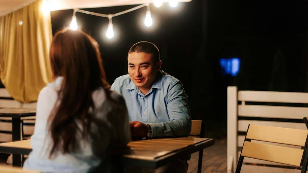 A man is sitting at an outdoor table at night, smiling at the woman sitting across from him.
