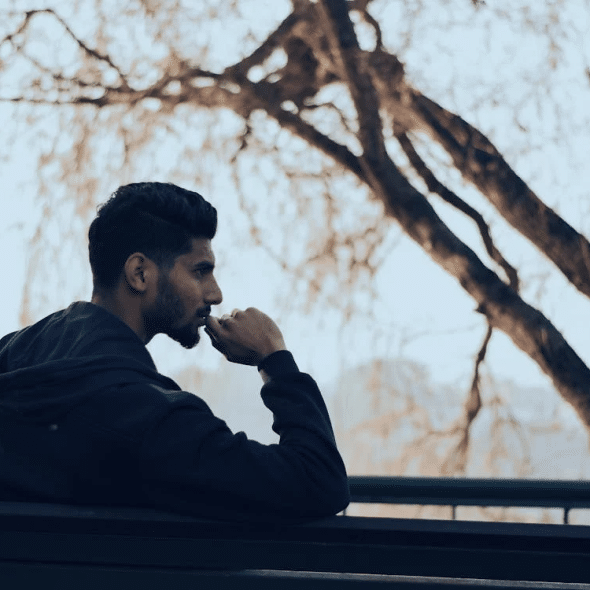 Man in Black Hoodie Sitting on Bench Near Green Trees