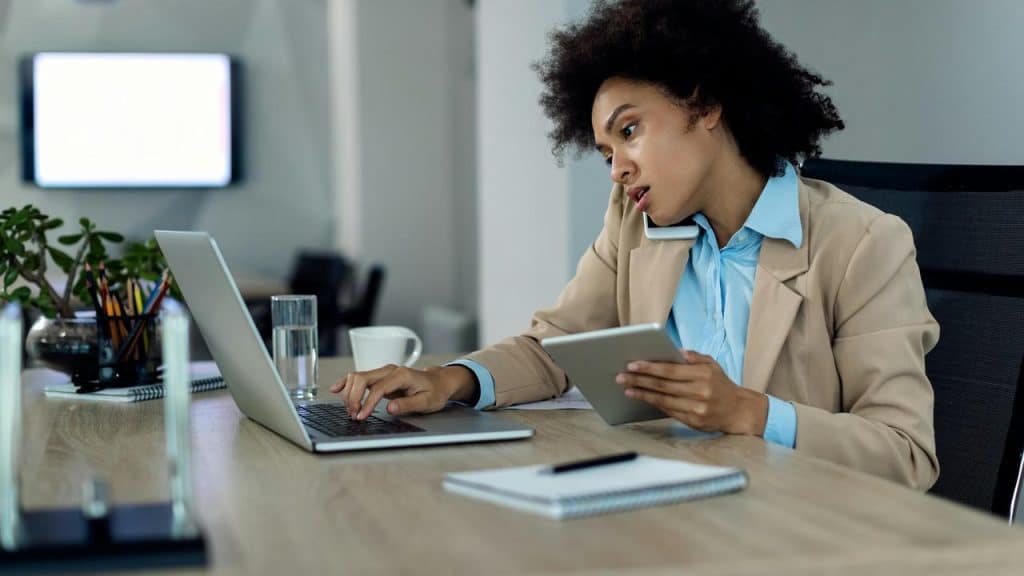 A woman talking on the phone while using a laptop and holding a tablet.
