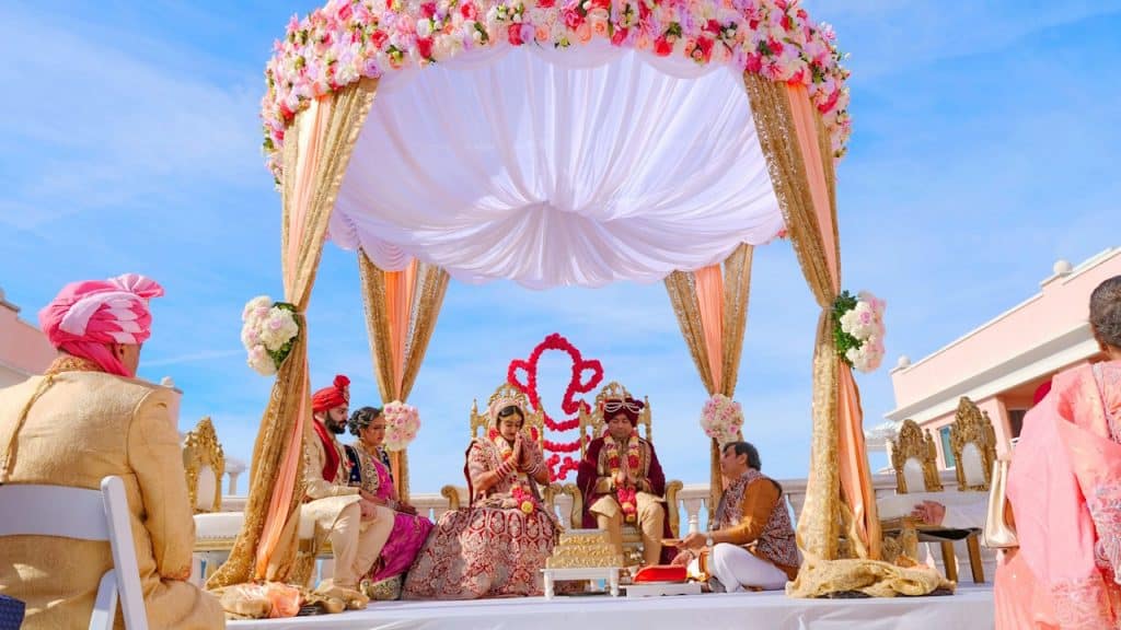 An Indian wedding ceremony outdoors under a pink and gold floral mandap on a bright, sunny day.