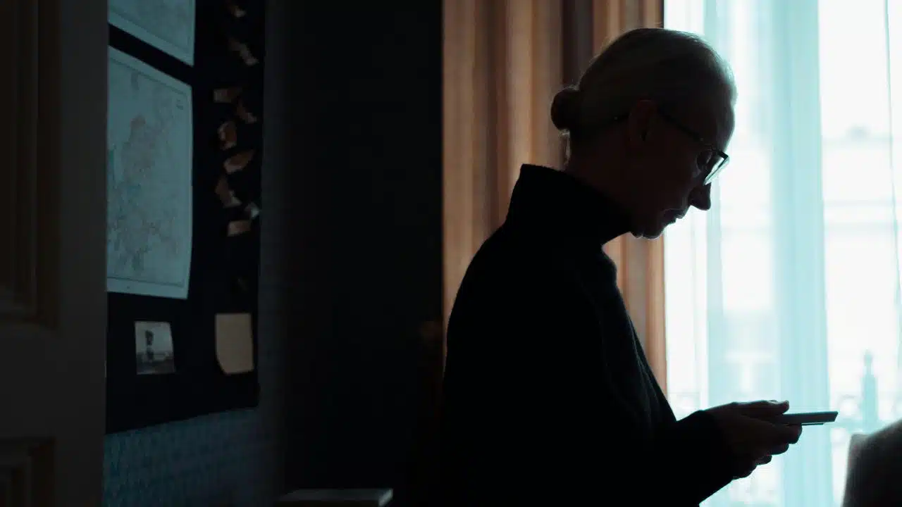 An older woman standing by a window while looking at her phone.