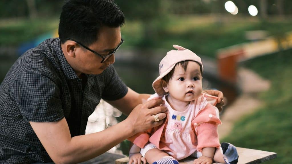 A man with glasses adjusts a baseball cap on a baby wearing a pink jacket outdoors.