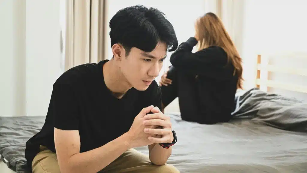 Man sits in the foreground with hands clasped, while a woman sits hunched behind him on a bed.