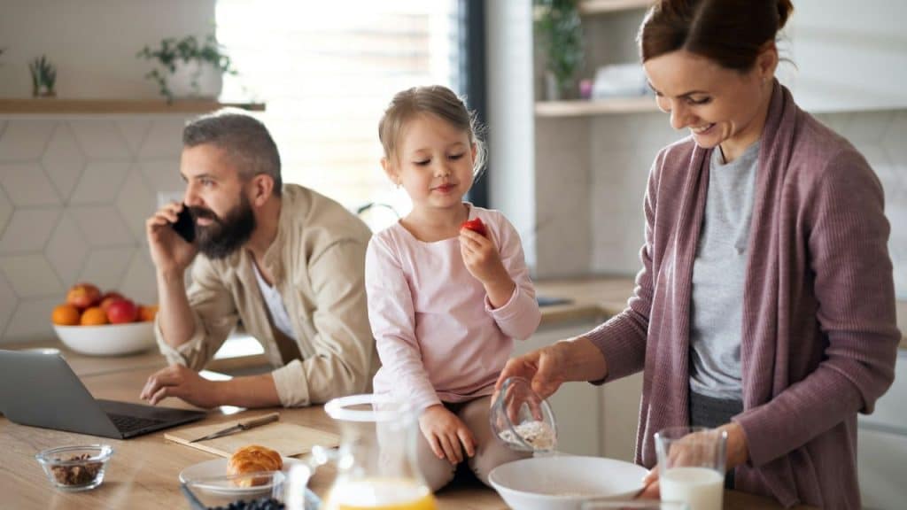 A family in a kitchen: a man on the phone, a girl eating, and a woman preparing food.