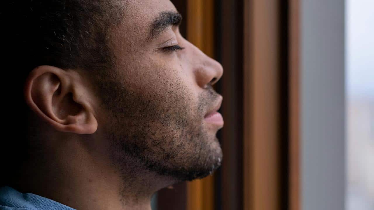 A close-up of a man resting his head against a window with his eyes closed.