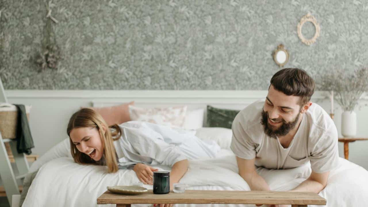 A couple laughing together while leaning over a low table in a cozy bedroom.