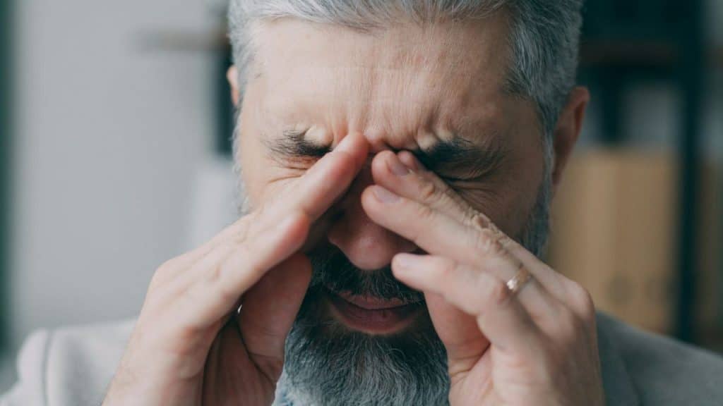 Close-up of a gray-bearded man rubbing the bridge of his nose in pain.