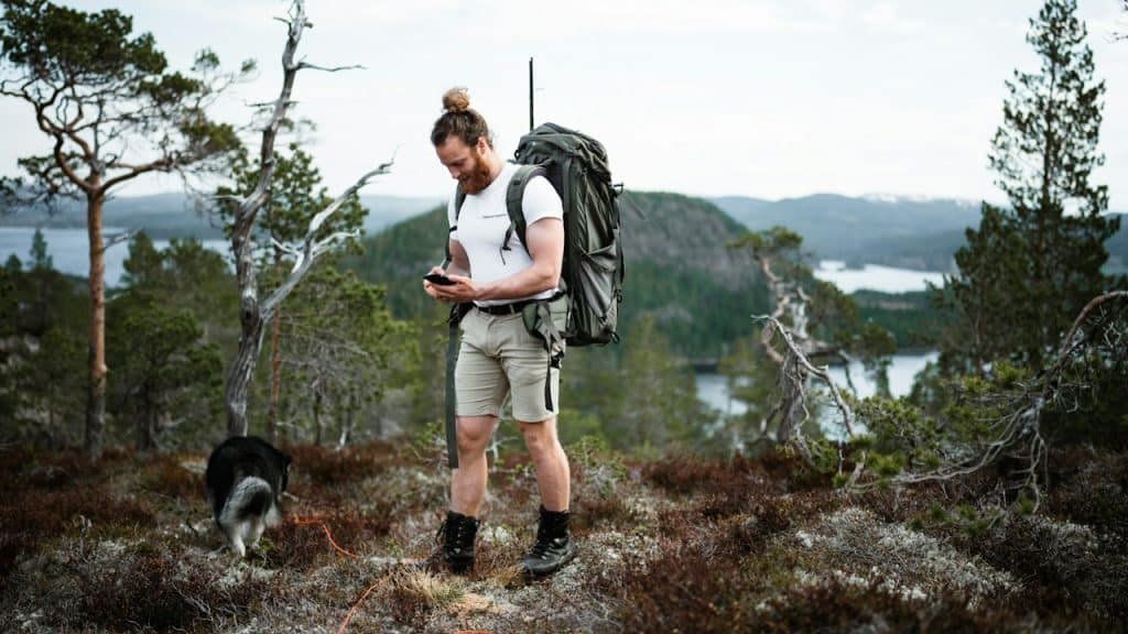 A bearded man with a hair bun trekking in the outdoors and looking down at his phone.