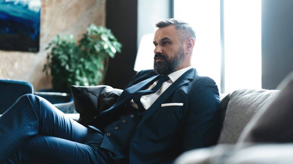 A bearded man wearing a suit sitting on a couch.