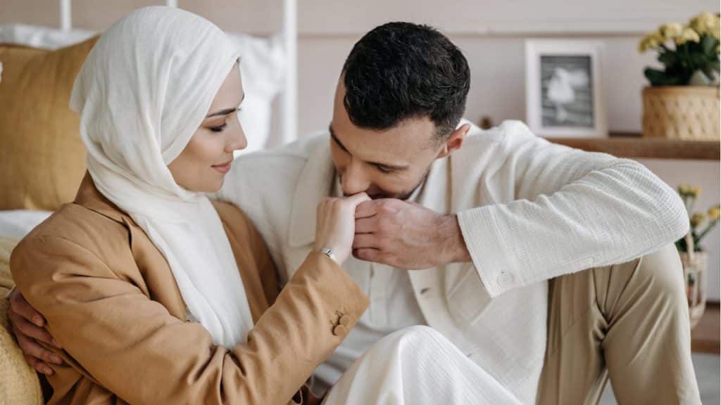 A man is sitting next to a woman wearing a hijab, kissing her hand, as they sit close together on the floor.