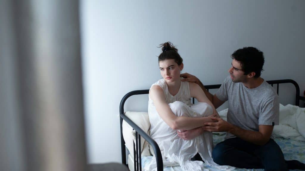 A man is sitting next to a distressed-looking woman, who is hugging her knees on a bed, as he tries to comfort her