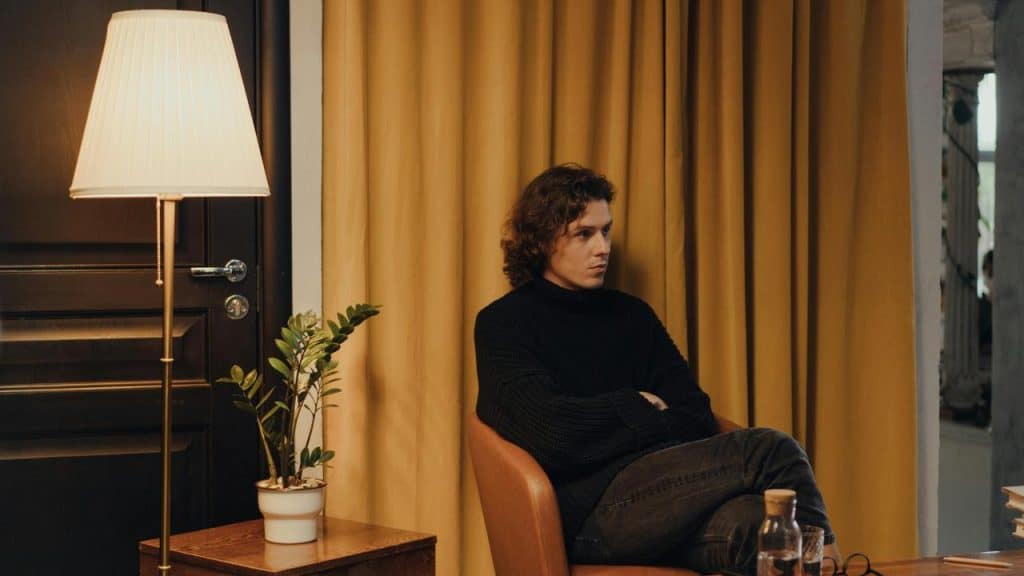 A man sitting in a chair with arms crossed, looking thoughtful beside a lamp and plant.