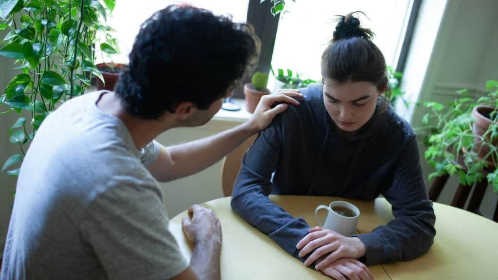 A man places a comforting hand on a woman's shoulder as she sits at a table.