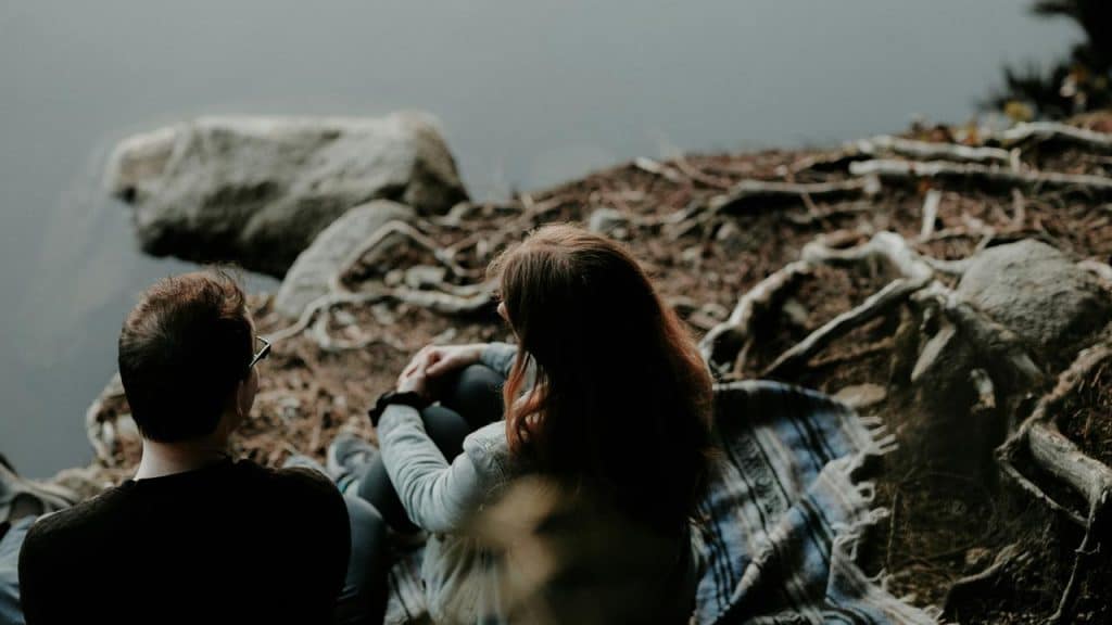 A man and a woman sitting together by a lakeshore, talking quietly.