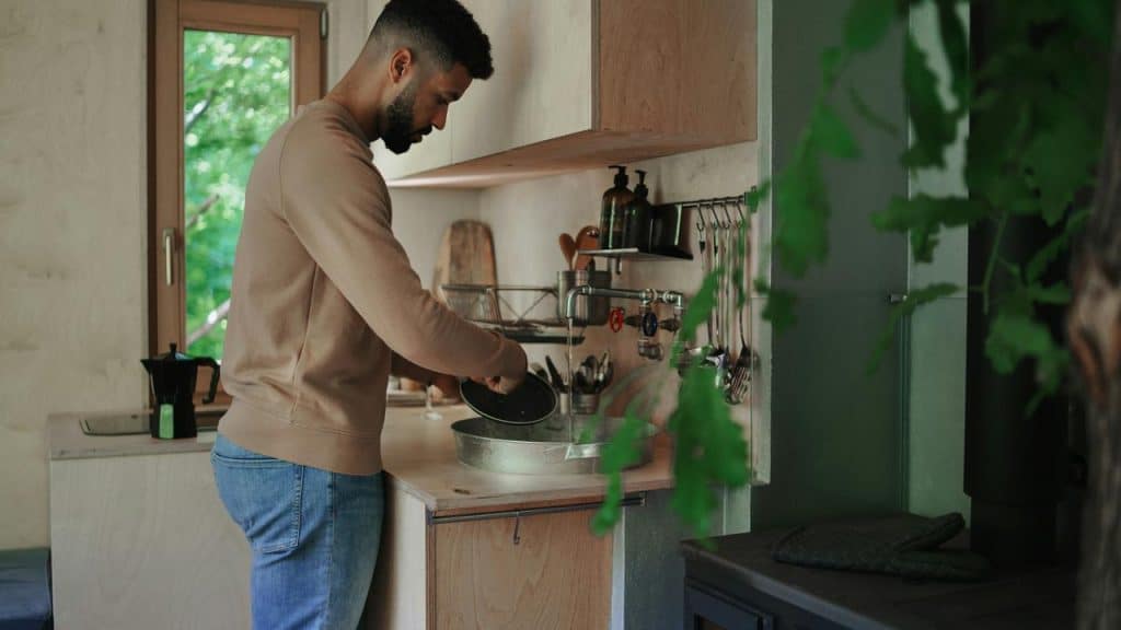 A man washing dishes in a small kitchen with a window overlooking greenery.