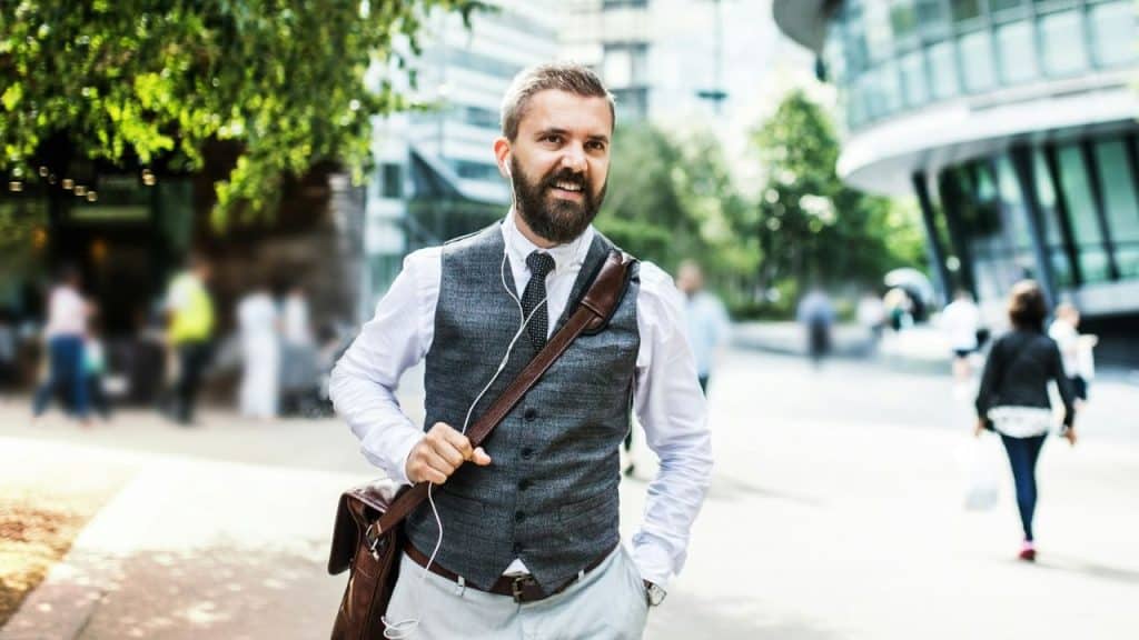 A bearded man in a vest and white shirt smiles with a leather bag on a busy sidewalk.