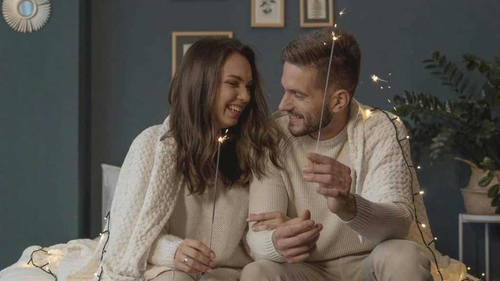 A smiling couple in cozy sweaters holds sparklers while sitting together.