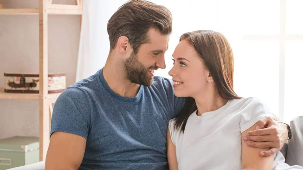 Smiling man with a beard embracing a woman, as they look into each other's eyes indoors.