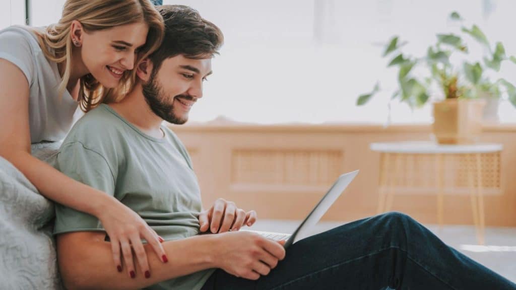 Smiling blonde woman embracing a man using a laptop while sitting on the floor.