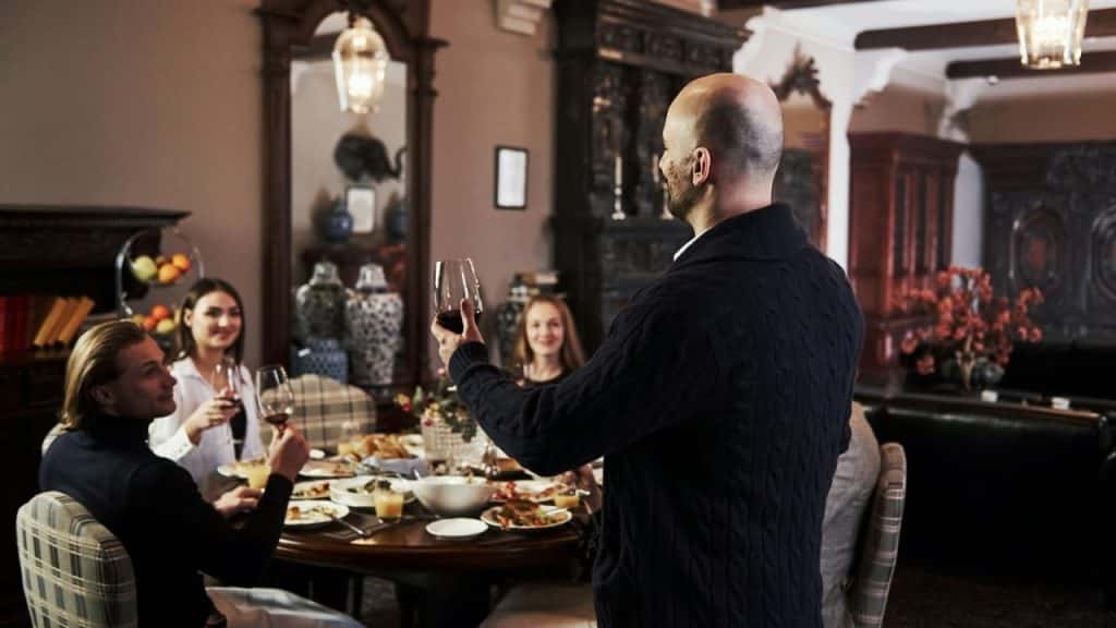 Bald man making a toast with a wine glass to people seated at a dinner table.