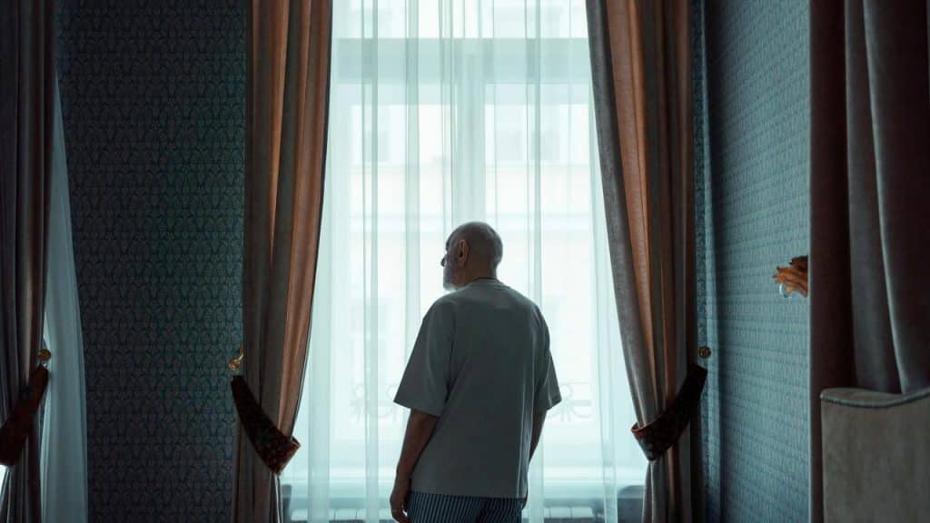 An elderly man standing by a window looking outside.