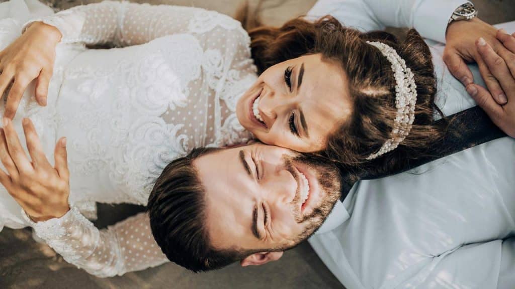 A bride and groom lie on the ground smiling joyfully, their faces close together.