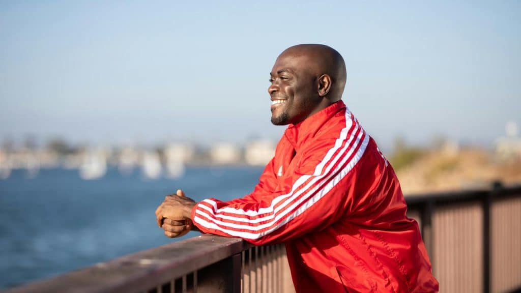 A man in a red jacket smiling while leaning on a railing by the water.