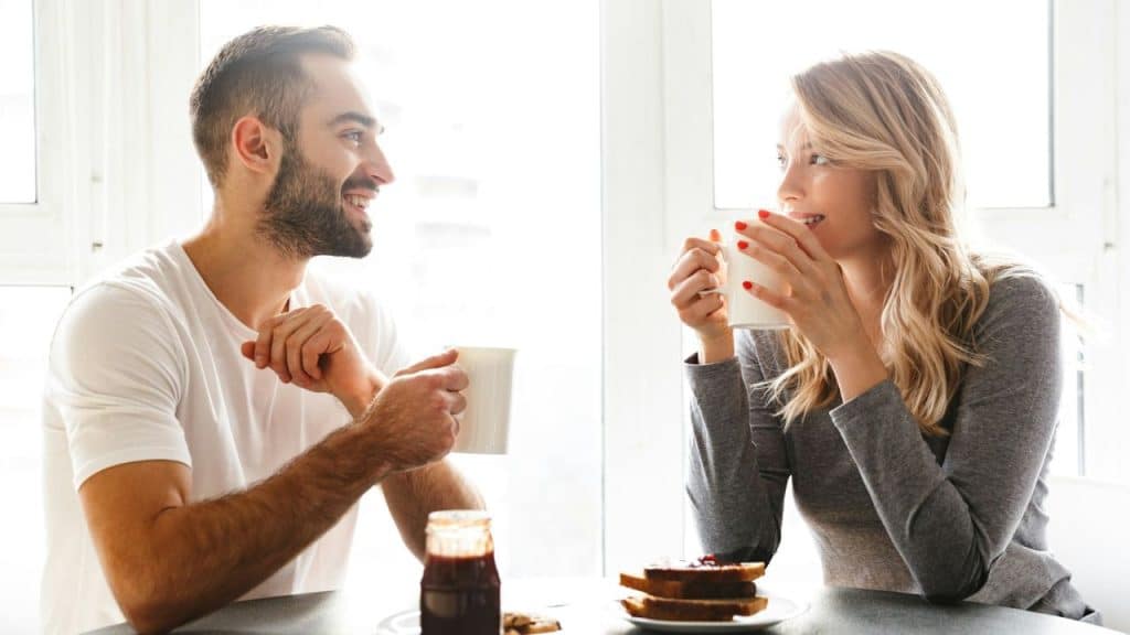 A happy couple smiles at each other while drinking from mugs at a sunlit breakfast table.