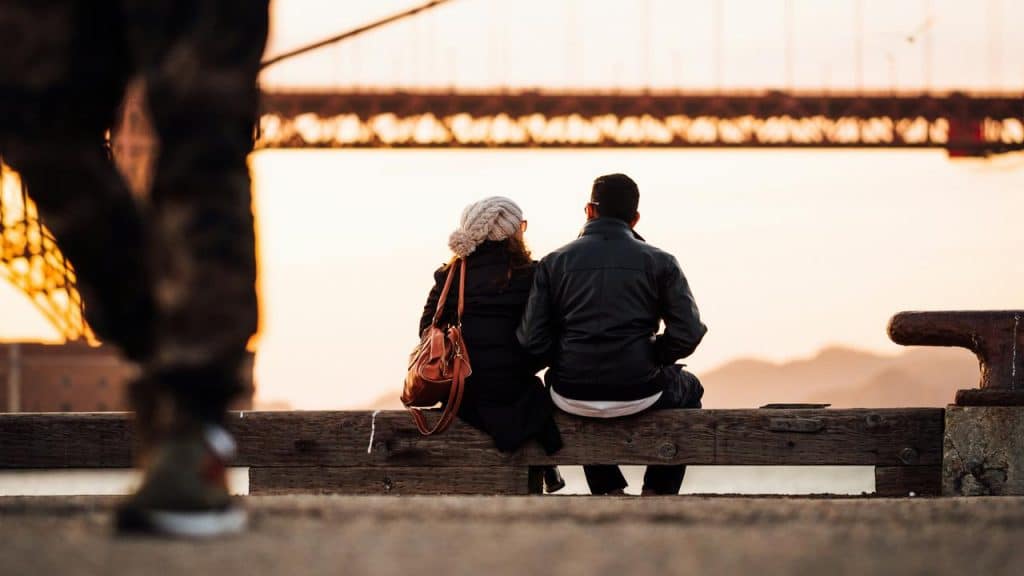 A couple sitting on a bench by the water, watching the sunset near a large bridge.