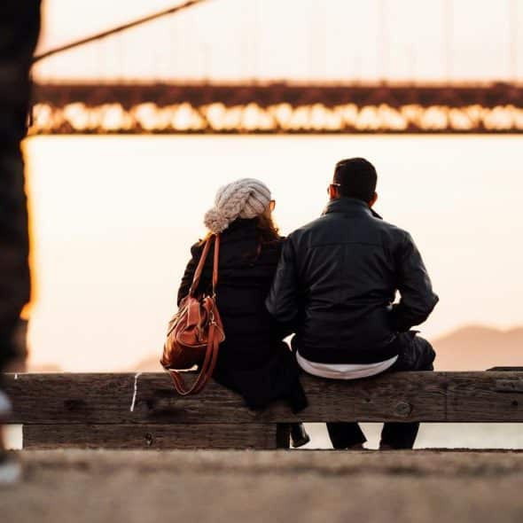 A couple sitting on a bench by the water, watching the sunset near a large bridge.