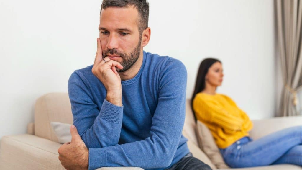 A serious man in a blue sweater sits with his chin resting on his hand; a woman is blurred in the background.