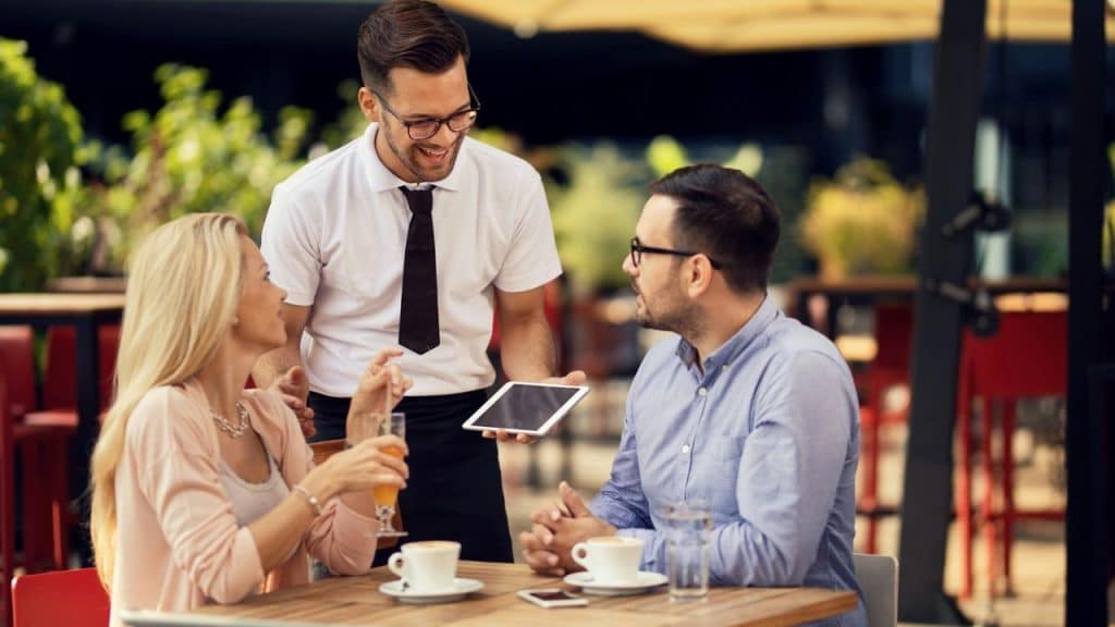A couple smiling and talking to a waiter holding a tablet at an outdoor cafe.
