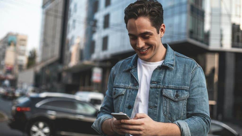 A man smiles while looking at his phone on a city street.