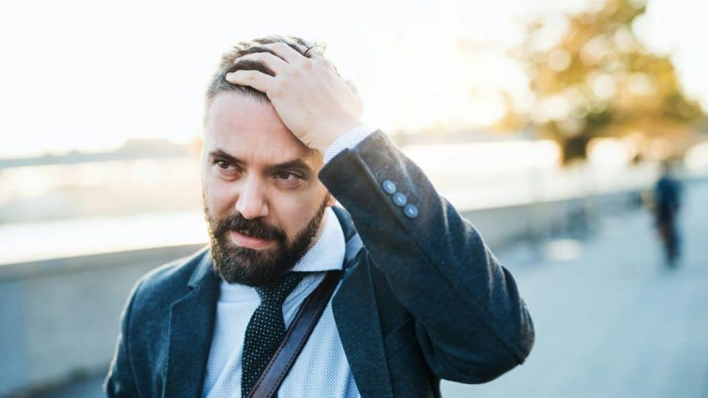 A bearded man outdoors runs his hand through his hair with a troubled expression.