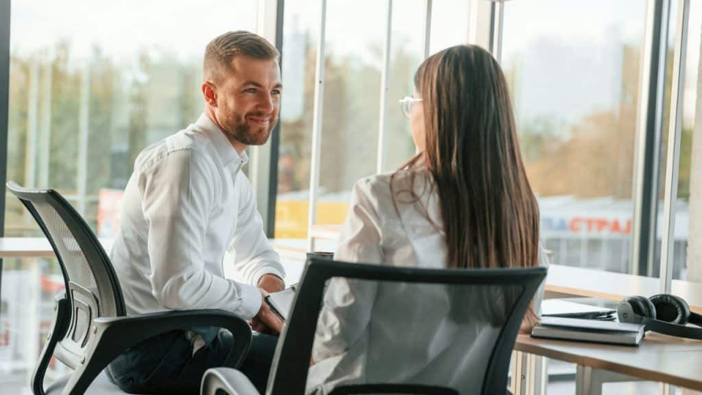 A smiling man in a white shirt looks at a woman across a table in an office.