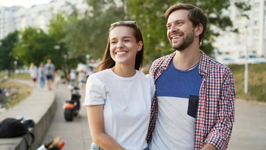 A smiling young couple, with the man's arm around the woman, stands outdoors on a sunny paved path.