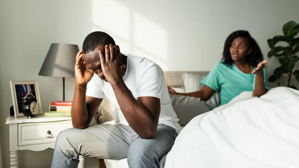 A distressed man sits on a bed with his head in his hands, while a woman argues behind him.