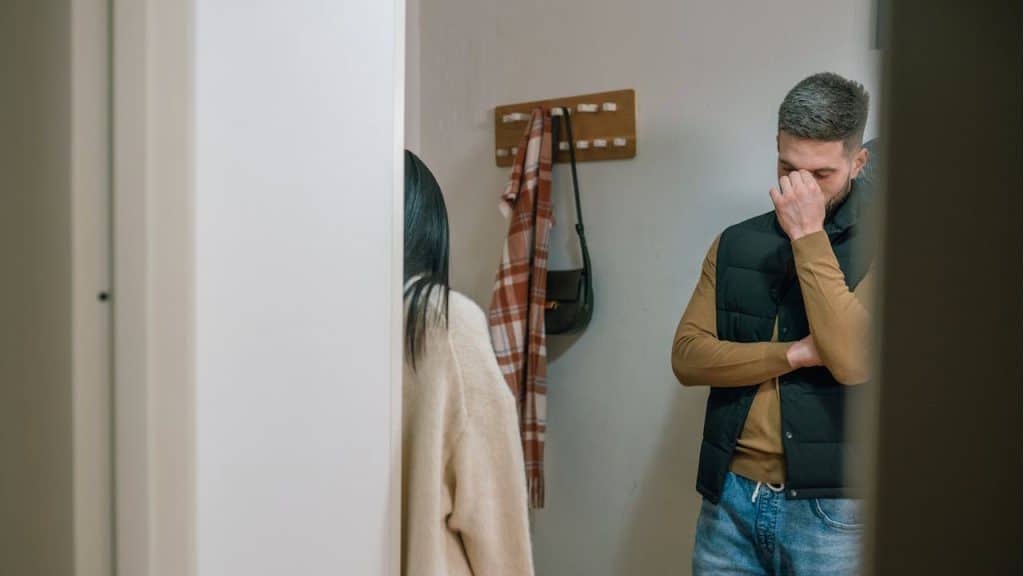 A man with a woman standing in a doorway with his hand covering his face, looking stressed or upset.