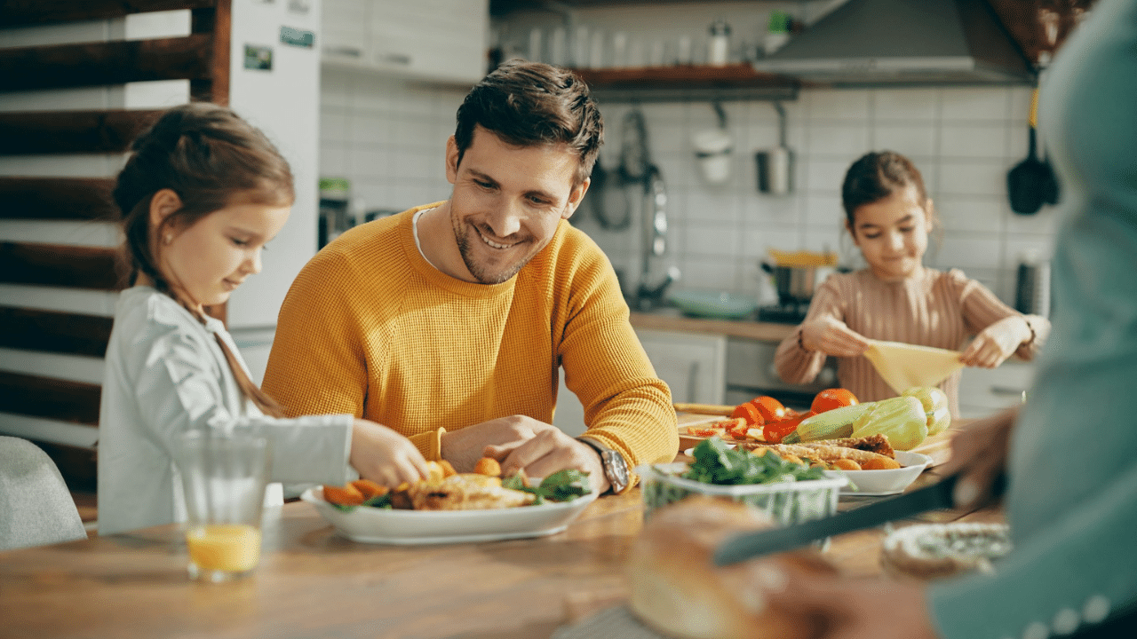 A smiling father and his two daughters eating a meal together in a bright kitchen.