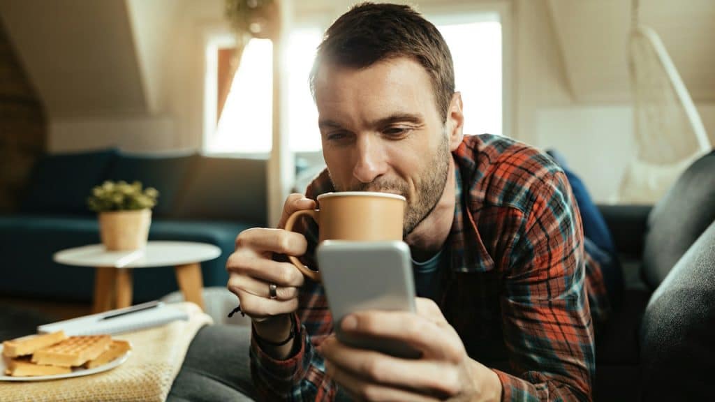 A relaxed man in a plaid shirt is lying on a couch, holding a coffee mug to his lips and looking at his smartphone.