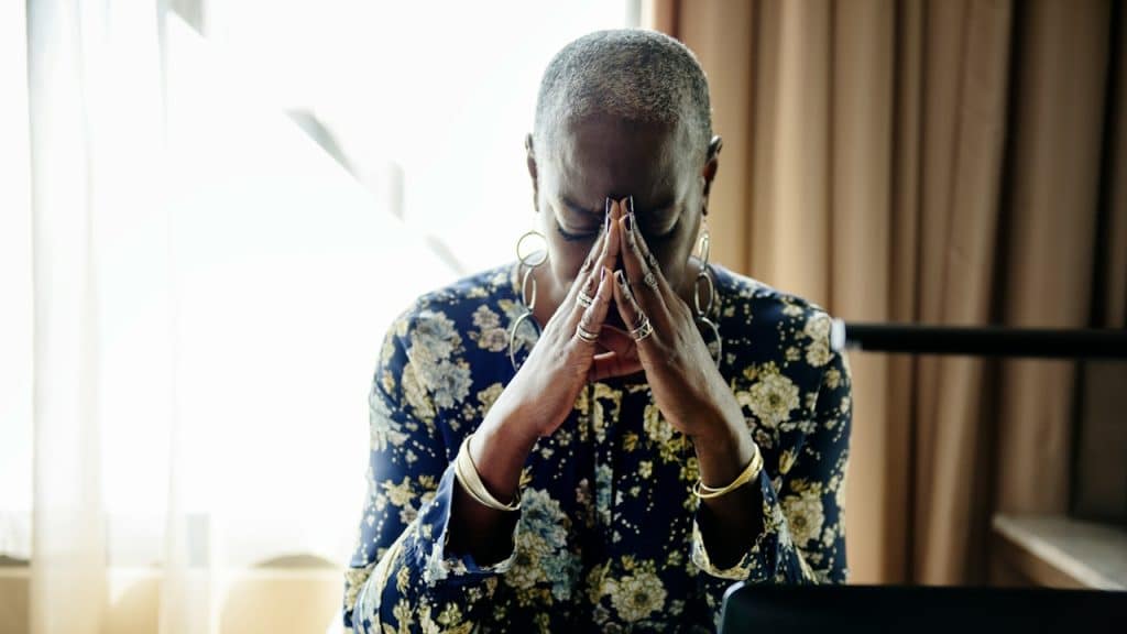 An older woman with earrings looking distraught and hiding her face in her hands.