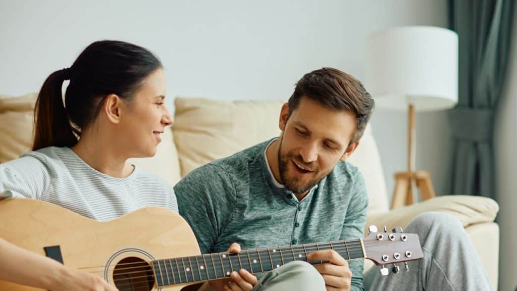 A woman learning to play acoustic guitar with the help of her husband at home.